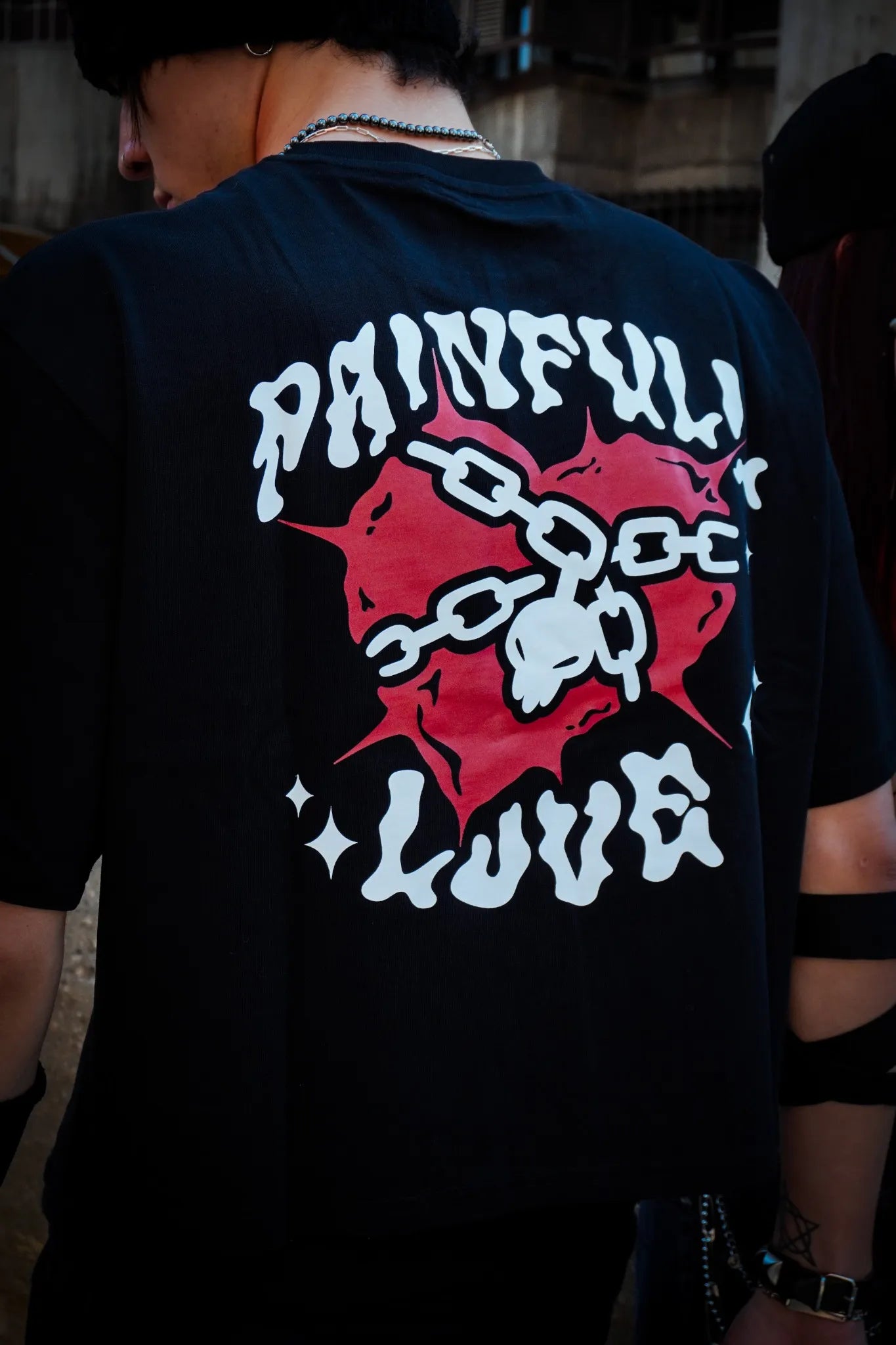 Persona de espaldas con camiseta “Painful Love” y diseño de calavera encadenada sobre fondo urbano